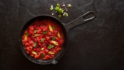 Cooking pasta penne with tomato sauce and basil served on iron cast pan. Black slate background, top view