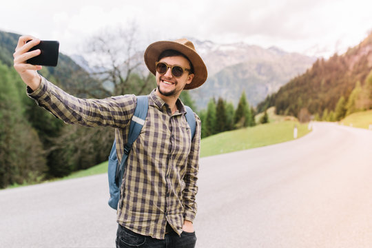 Smiling man in brown hat standing with hand in pocket and making selfie while catches the car on road. Stylish laughing guy wearing sunglasses taking photo of himself during vacation trip.