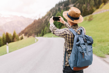 Male traveler in trendy checkered shirt making photo of amazig landscape with forest and mountains. Outdoor portrait of man in hat with backpack standing on the road with smartphone during trip.