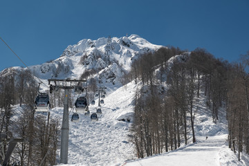 Snow-capped mountains of the ski resort Krasnaya Polyana in Sochi on which the cable car with tourists. Clear blue sky in the mountain forest. Tourist season of skiing and snowboarding