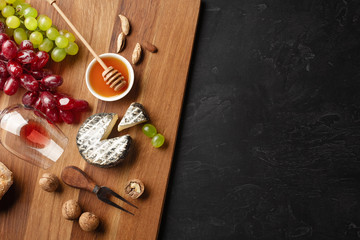 Cheese head, bunch of grapes, honey, nuts and wineglass on wooden board and black background