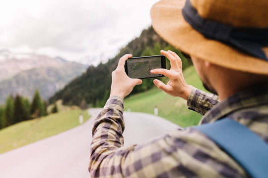 Close-up portrait from back of traveler searching address in online map during trip in italian mountains. Young man in checkered shirt and hat decorated with ribbon looking at his route holding phone.