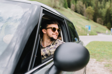 Handsome man in good mood drives a car and enjoying the fine weather on vacation. Smiling traveler wearing trendy sunglasses looking away on amazing nature view during trip around Europe.