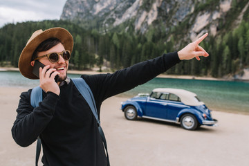 Brunette man wears sunglasses and black sweatshirt speaking on phone during walk along river shore and pointing finger on something interesting. Outdoor portrait of laughing tourist looks on mountain.