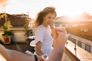 Outdoor portrait of happy touched young woman in white shirt with male hand holding ring on foreground. Attractive cute girl looking with love to camera got an offer to get married.