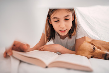 Girl lying on the bed under blanket with her dog and reading book