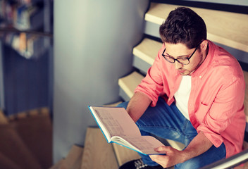 education, high school, university, learning and people concept - student boy or young man reading book sitting on stairs at library