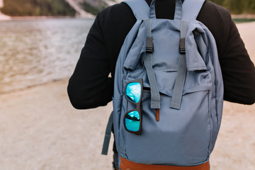 Man wearing cotton black sweater standing in front of lake with stylish travel bag and sunglasses. Outdoor portrait of guy carrying big blue backpack walking near river with rucksack on foreground.