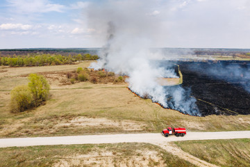 Aerial view of red fire truck on a country road near big fire in the field with burnt land and...