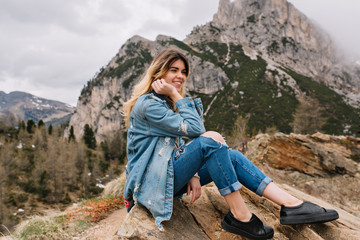 Naklejka na ściany i meble Dreamy fair-haired girl wearing denim attire resting on the rock after hard climbing and posing touching her face. Attractive stylish young woman with charming smile sitting on stone on mountain view.
