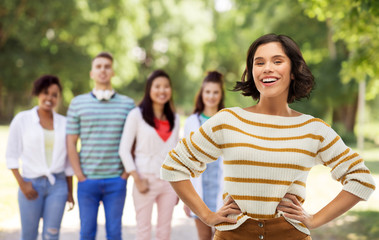Obraz premium people concept - happy smiling young woman in striped pullover with hands on hips over group of friends in summer park background