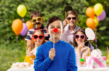 red nose day and photo booth concept - smiling boy in hoodie with clown nose over group of friends at birthday party in summer park background