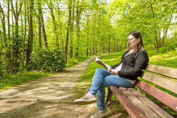 Fototapeta premium Woman stands reading sitting on a bench of a public park enjoying nature
