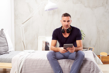 Caucasian man in forties sitting on bed in bedroom and using tablet.