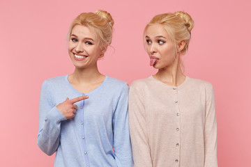 One of beautiful young blonde twins is fooling around and showing her tongue, her sister smiles broadly and pointing at her isolated over pink background. - obrazy, fototapety, plakaty