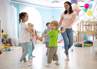 Group of happy children standing in circle holding hands, playing with their teacher in daycare