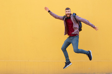 Fototapeta na wymiar Happy smiling young attractive ginger bearded man jumping, wearing in basic clothes with backpack. Looking at the camera with wide open mouth over a yellow wall with copy space.