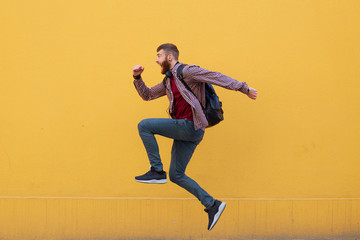 Young attractive ginger bearded man jumping, very fast running, hurry to the store for sale, wearing in basic clothes with backpack. Isolated over yellow wall with copy space.
