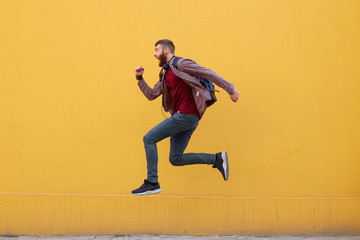 Fototapeta na wymiar Young attractive ginger bearded man jumping, very fast running, running away from hateful work, wearing in basic clothes with backpack. Isolated over yellow wall with copy space.