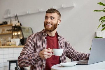 Photo of happy smiling attractive ginger bearded man working at a laptop, sitting in a cafe,...