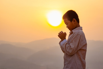 Fototapeta premium little boy praying to the GOD while holding a crucifix symbol with bright sunbeam on the sky