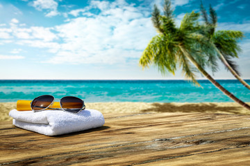 Obraz na płótnie Canvas Wooden table background of free space for your decoration. White towel background with sunglasses and beach landcsape with palms and ocean. Blue sky with sun light. Summer time on beach. 