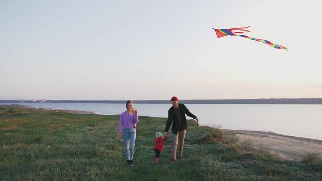 Happy family walking with flying kite on field near seashore during sunset, slow motion