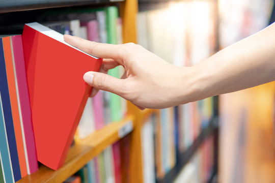 Bestseller publishing concept. Male hand choosing and picking red book from wooden bookshelf in bookstore. Education research in university public library.
