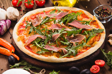 Pizza with bacon and arugula on a dark wooden background with ingredients around (close).