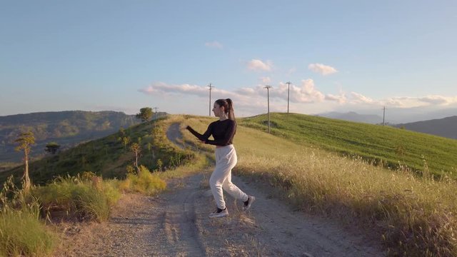 Giovane ragazza con maglia nera e tuta grigia balla con musica allegramente in una strada sterrata, in cima a una collina in campagna. Tra la natura. Tramonto sullo sfondo.