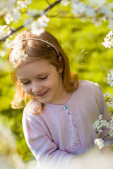 Portrait of cute and pretty little girl looking down in blooming trees. Adorable little girl smiling in spring garden on sunny day. Child and flowers. Childhood & natural tenderness. Children's day. 