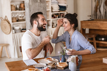 Image of modern brunette couple eating breakfast together while sitting at table at home - obrazy, fototapety, plakaty