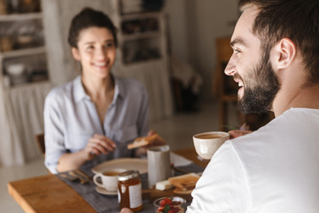 Image of positive brunette couple eating together at table while having breakfast in apartment
