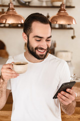 Portrait of smiling brunette man 20s drinking coffee and using smartphone at home