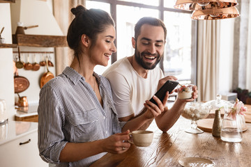 Photo of european brunette couple drinking coffee and using cell phone during breakfast in kitchen...