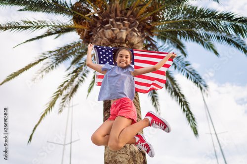 Happy child teenager girl jumping is waving American USA flag at background of palm tree