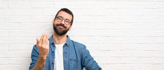 Handsome man with beard over white brick wall inviting to come with hand. Happy that you came - obrazy, fototapety, plakaty