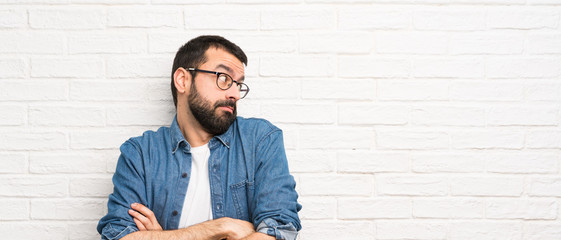 Handsome man with beard over white brick wall making doubts gesture while lifting the shoulders