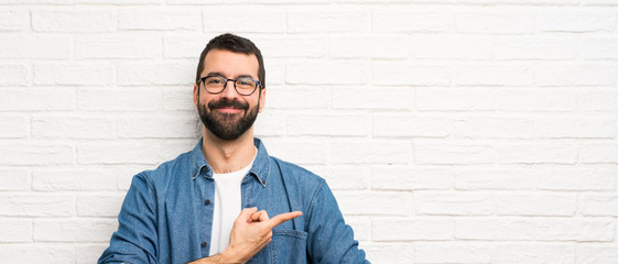 Handsome man with beard over white brick wall pointing to the side to present a product