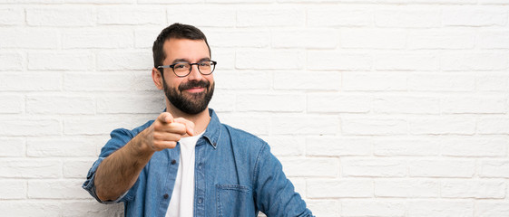 Handsome man with beard over white brick wall points finger at you with a confident expression