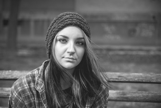 Homeless girl, Young beautiful girl sitting alone outdoors on wooden bench on the street with hat and shirt feeling anxious and depressed after she became a homeless person black and white moody image
