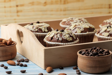 Delicious, sweet chocolate muffins, with almond petals in a wooden tray next to grains of coffee on a blue wooden table.