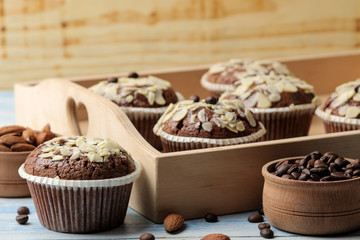 Delicious, sweet chocolate muffins, with almond petals in a wooden tray next to grains of coffee on a blue wooden table.