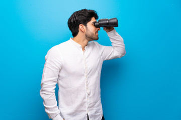 Young man over isolated blue wall and looking in the distance with binoculars