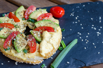 Bagel with hummus, avocado slices, spices, green onion, tomatoes cherry and sesame seeds on the black stone desk. Close up.