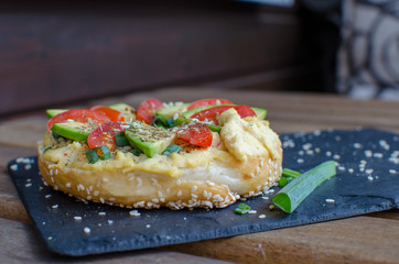 Bagel with hummus, avocado slices, spices, green onion, tomatoes cherry and sesame seeds on the black stone desk. Close up.