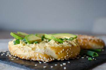 Bagel with hummus, avocado slices, spices, green onion, and sesame seeds on the black stone desk. Close up. Side view