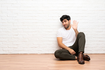 Young man sitting on the floor saluting with hand with happy expression