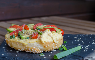 Bagel with hummus, avocado slices, spices, green onion, tomatoes cherry and sesame seeds on the black stone desk. Close up.