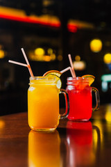 Two refreshing cold summer lemonades in glass jars, orange and red, served with ice, orange and lime slices. Shot in a bar using studio lights, reflections in a bar stand.